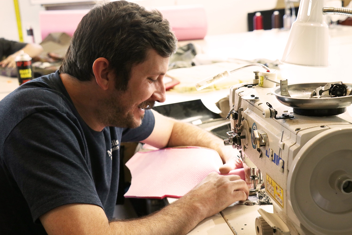 There's a man sewing upholstery for a couch. The man is part of the furniture team and is smiling.