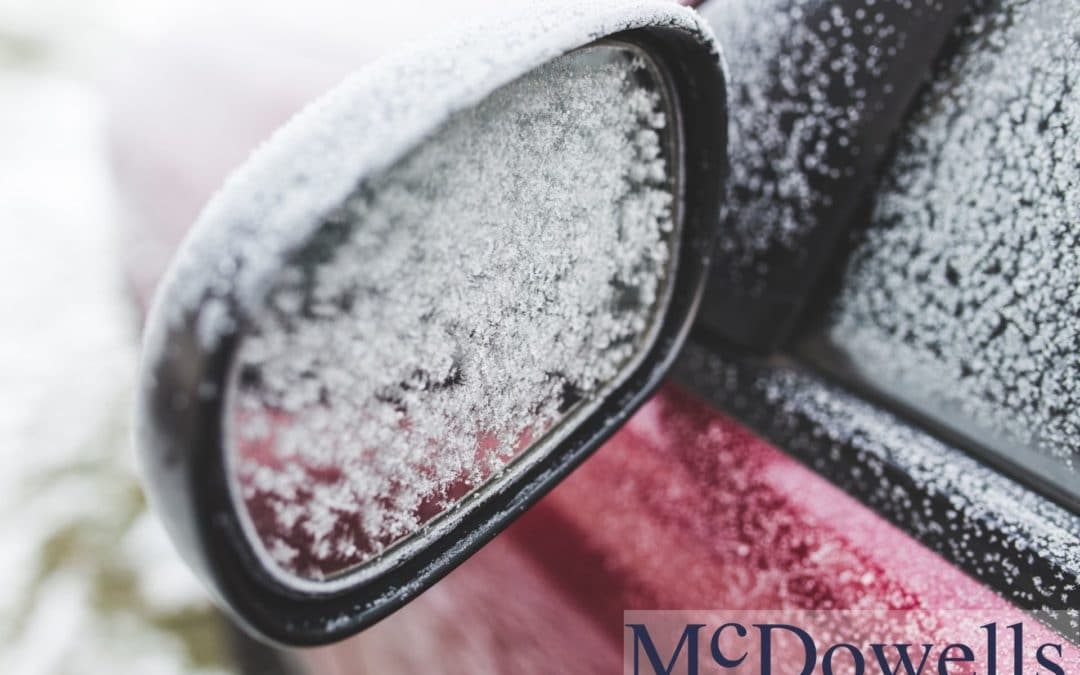 The driver’s side-view mirror of a car. The mirror is frosted over from the winter weather.